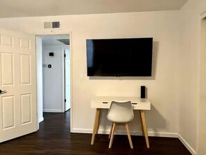 Simple, well-lit room with a desk, chair, and flat-screen TV.
