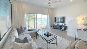 A spacious living room with a large sofa, a coffee table, and a TV. There is a balcony with a view of the ocean.