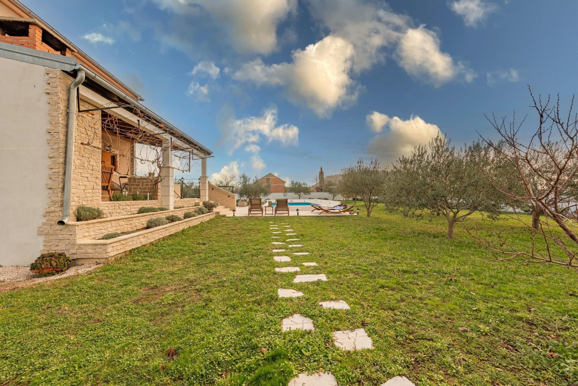 A house with a stone exterior, a garden with stepping stones, and a pool in the background.
