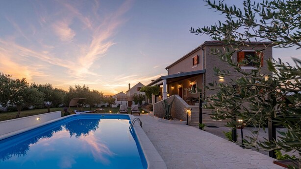 Una piscina con solárium de piedra, rodeada de un jardín ajardinado y un edificio con fachada de piedra.