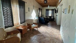 Amplia sala de estar con techo alto, vigas de madera y ventilador de techo. Se observan muebles de mimbre, un sofá y una mesa de comedor. El piso está cubierto de baldosas y hay grandes ventanas con cortinas.