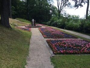 Uma trilha no jardim com uma fonte central, rodeada por canteiros de flores.