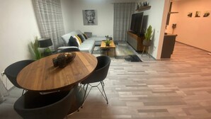 Een moderne woonkamer met een eethoek, een grijze bank, een televisie en decoratieve planten.