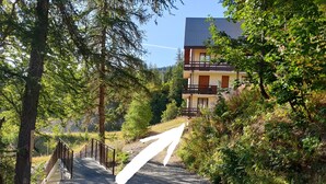 Ein Kiesweg führt zu einem zweistöckigen Gebäude mit Balkon, umgeben von dichten Bäumen und Grün.