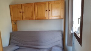 Ein kleines Zimmer mit einem Einzelbett, Holzschränken darüber und einem Fenster mit Vorhängen.
