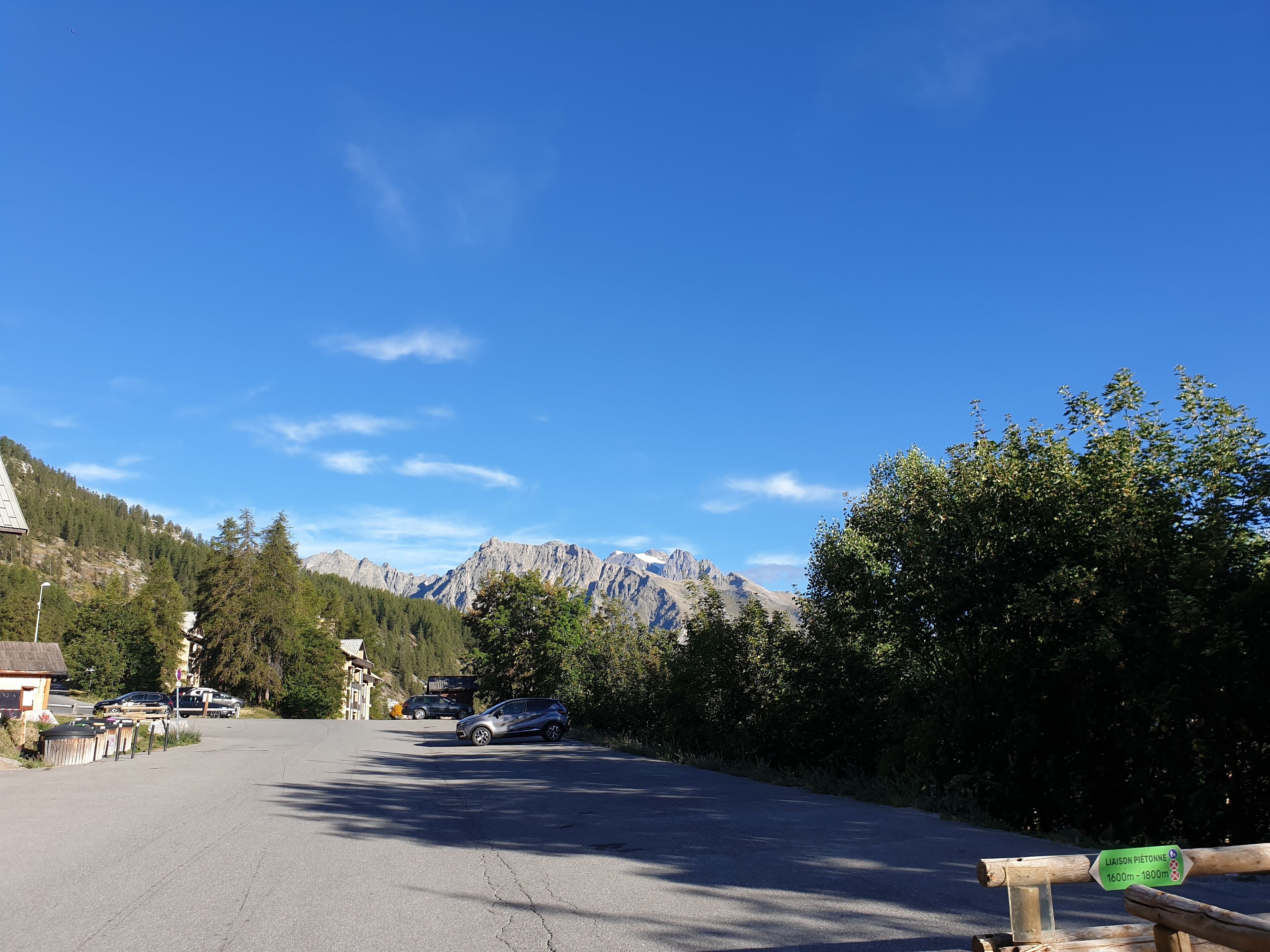 Una strada con auto parcheggiate sul lato, un segnale stradale e una catena montuosa sullo sfondo.