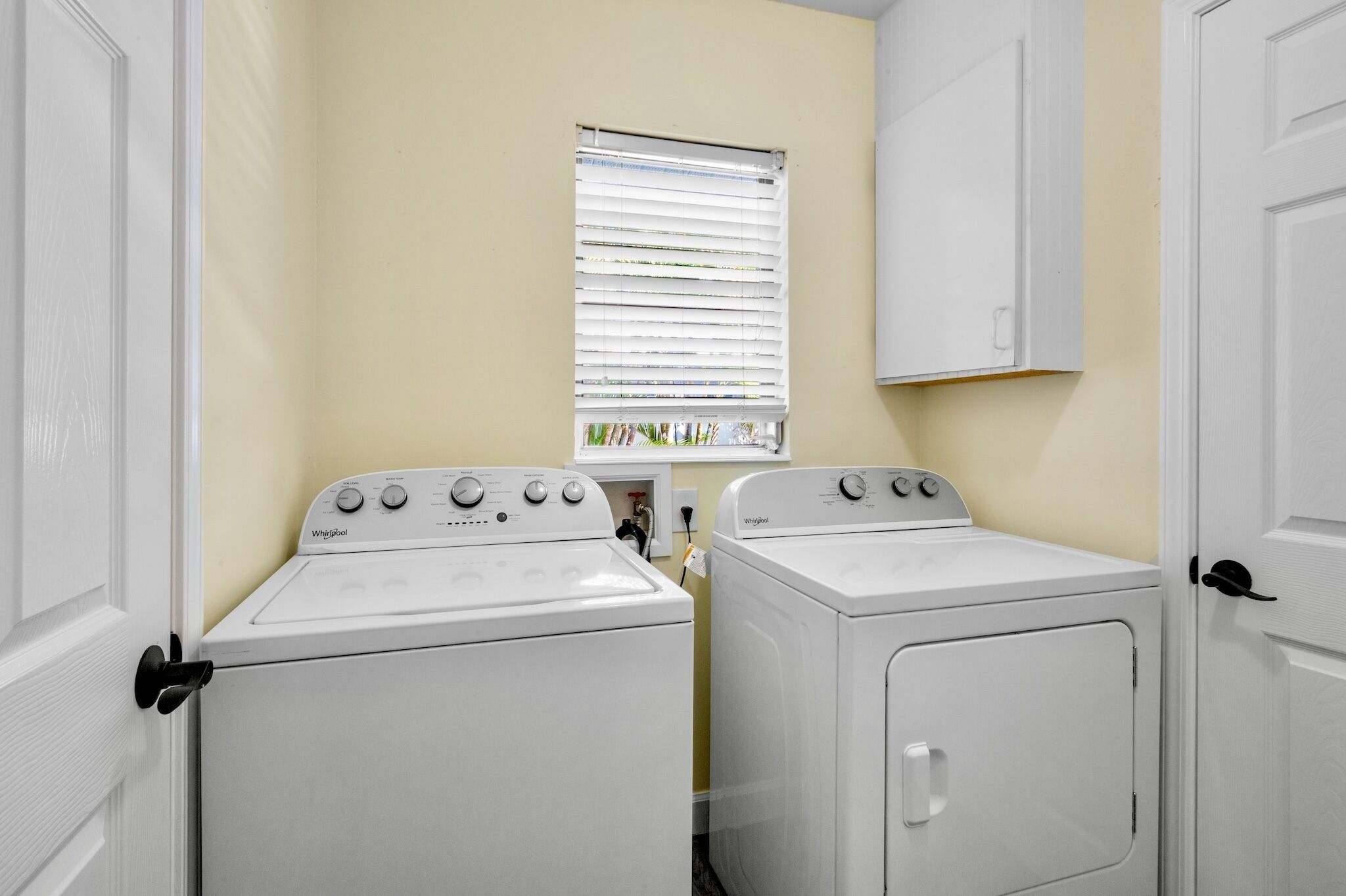 Uma lavanderia com duas máquinas de lavar e secar Whirlpool, uma janela com persianas e um armário branco acima das secadoras.