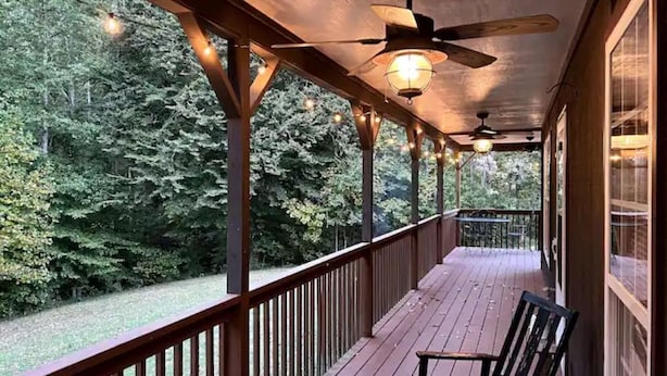 Una terraza de madera con ventilador de techo y luces colgantes, con vista a una zona boscosa.