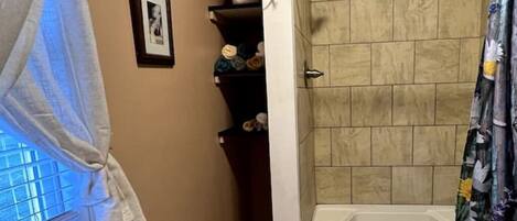 A bathroom with a shower, a window with a curtain, and shelves with toiletries.