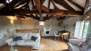A rustic room with exposed wooden beams, a white sofa, a hanging chair, a small table, and a window with a view of greenery.
