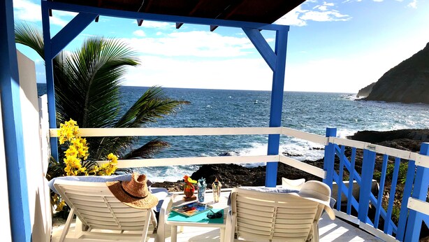 Un espacio de estar al aire libre cubierto, con sillas blancas y una mesa, con vistas a un litoral rocoso y al océano.