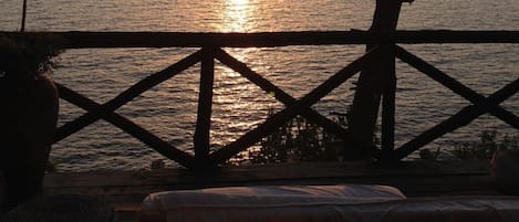 A sunset view from a balcony with a railing, overlooking a calm sea and distant land.