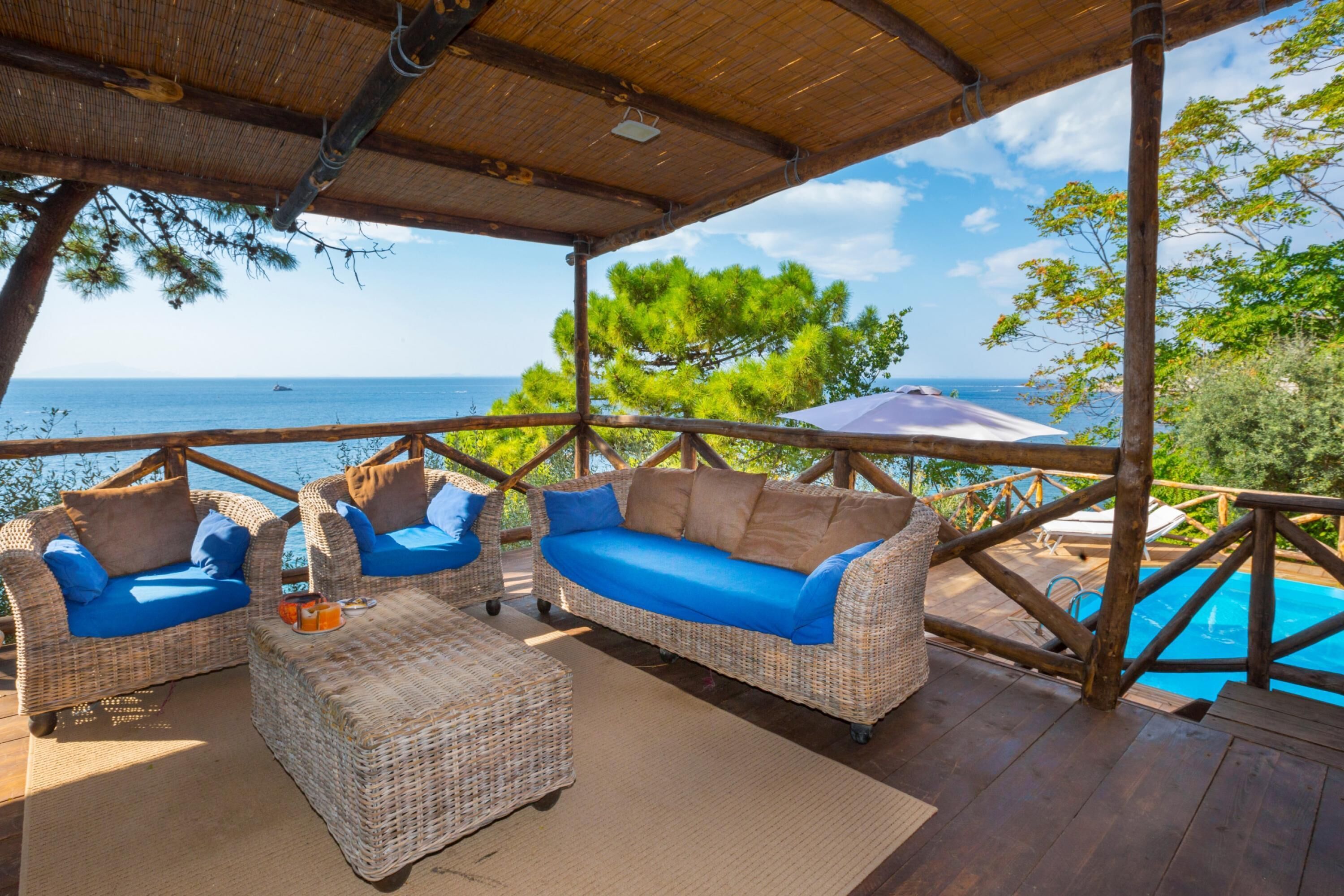 Uma área de estar coberta ao ar livre, com móveis de vime, piscina e vista para o mar.