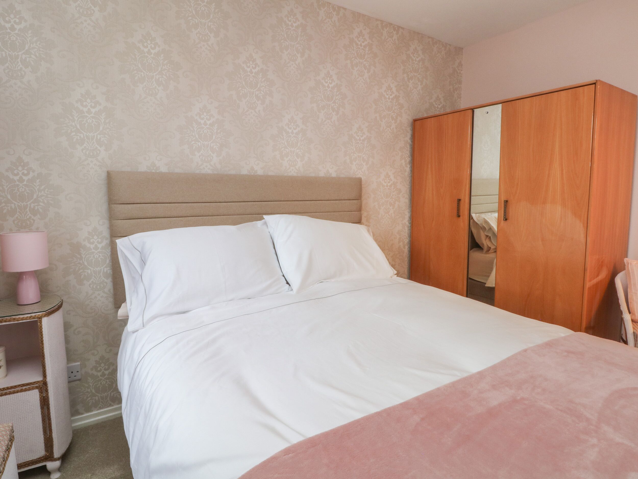 Ein ordentlich bezogenes Bett mit weißer Bettwäsche, ein Holzschrank und ein kleiner Nachttisch mit einer rosa Lampe.