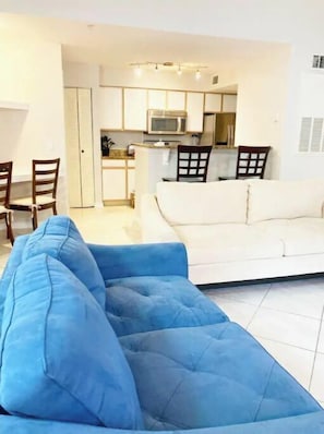 Uma sala de estar com um sofá azul, um sofá branco e uma área de cozinha ao fundo.