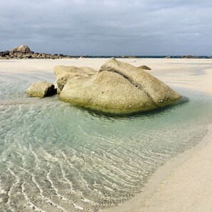 A clear beach with large rocks in shallow water and a distant rocky shoreline.
