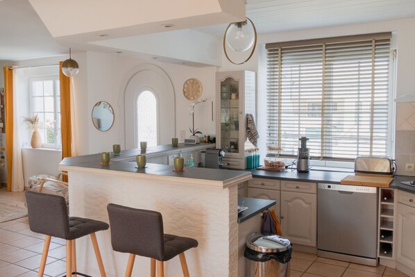 A modern kitchen with a central island, bar stools, a dishwasher, and a window with blinds.
