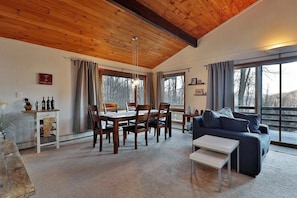 A cozy living room with a wooden ceiling, a dining area with a table and chairs, a sofa, and large windows offering a view of trees.