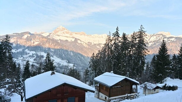 Une cabane en bois dont le toit est recouvert de neige, entourée d’arbres et de montagnes également enneigés, qui se dressent à l’arrière-plan.