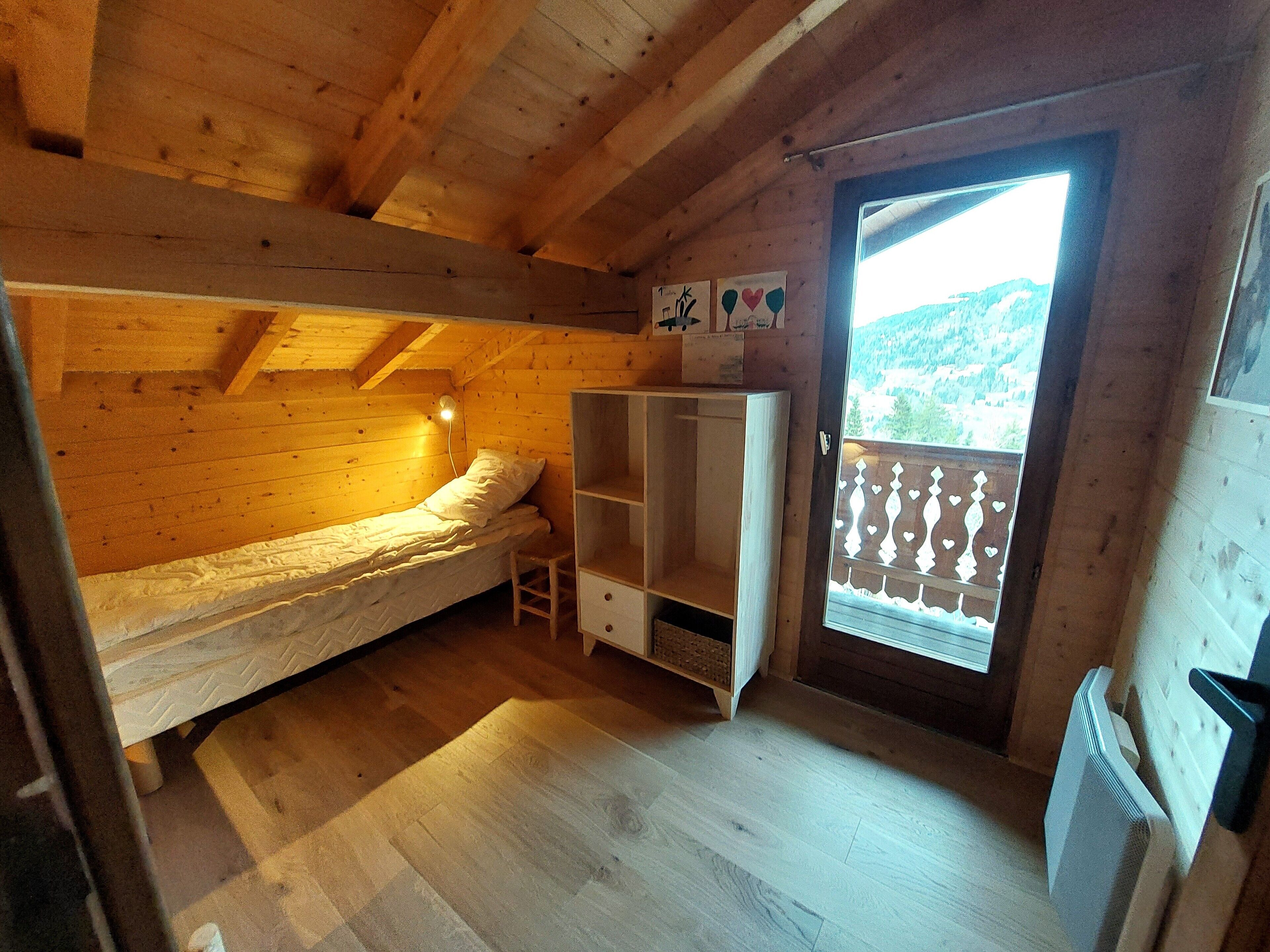 A small wooden cabin room with a bunk bed, a wooden wardrobe, and a balcony with a view of the mountains.