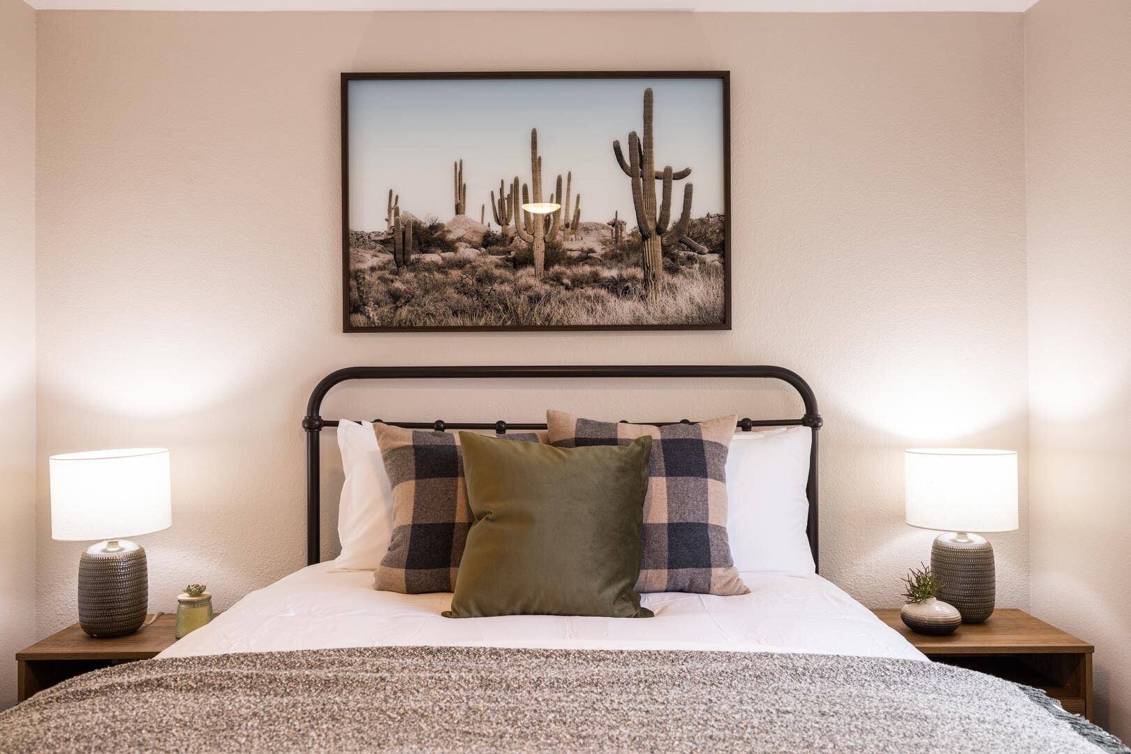 Een slaapkamer met een bed, twee bedlampen, een lijst met cactussen en een geruit kussen.