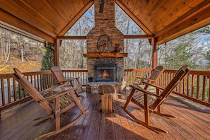 Eine Holzterrasse mit einem Kamin aus Stein, zwei Schaukelstühlen und Blick auf den Wald.