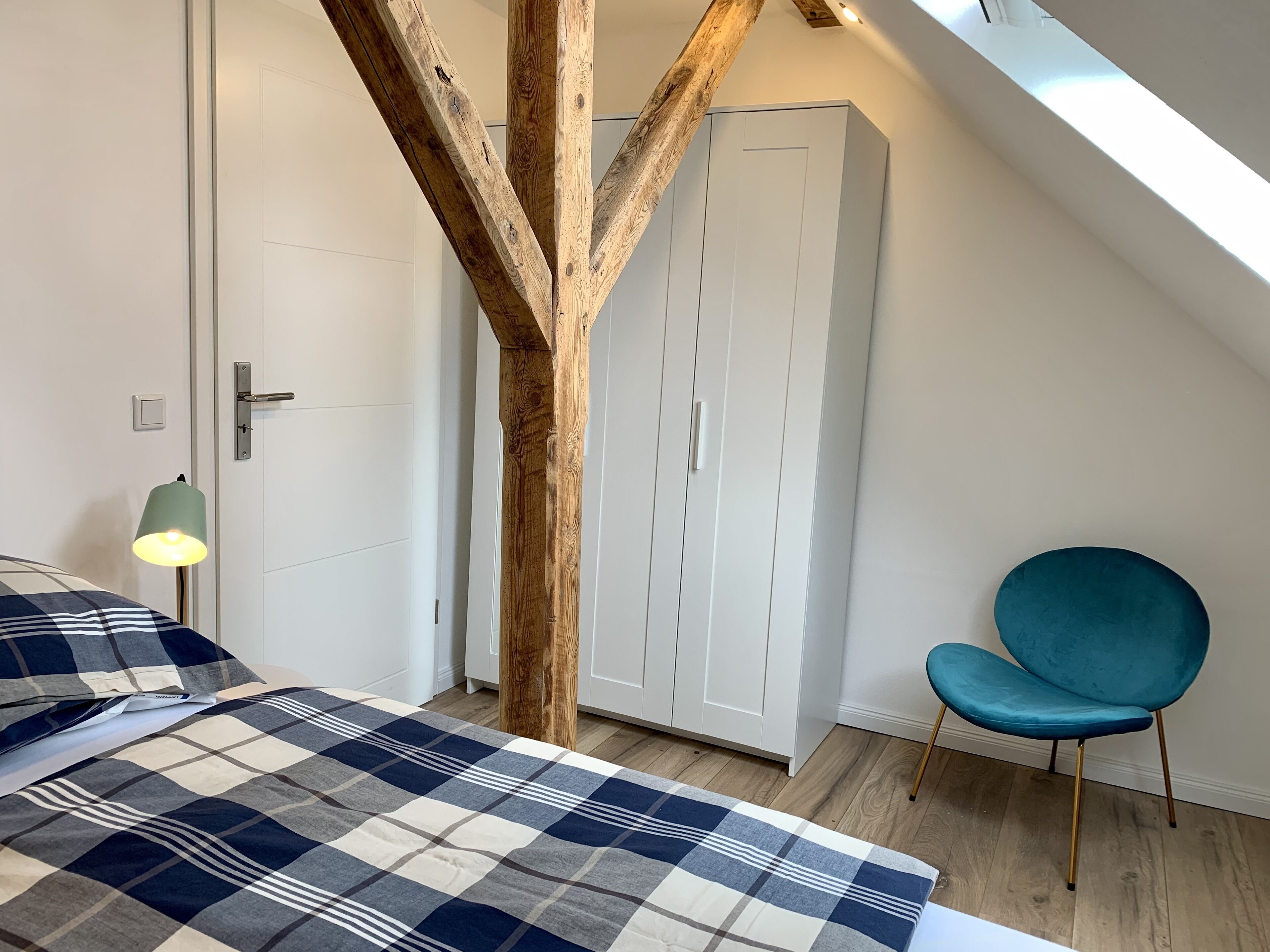 A bedroom with a checkered bedspread, a wooden beam structure, a white wardrobe, a teal chair, and a bedside lamp.