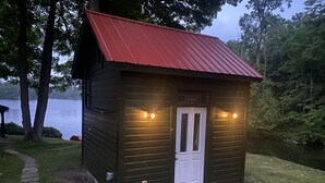 A small, dark wooden cabin with a red roof, a white door, and two exterior lights. - Cozy Lakefront Getaway in South Frontenac, Rustic & Elegant, Highspeed Internet (Battersea)