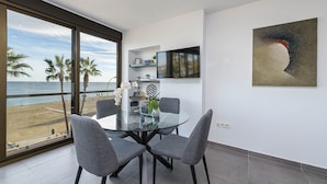 A modern dining area with a glass table and grey chairs, overlooking a beach with palm trees.