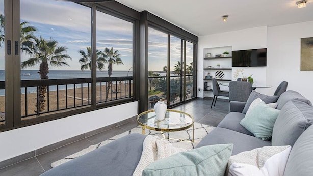 A modern living room with a large balcony overlooking a beach, a dining area, and a flat-screen TV mounted on the wall.