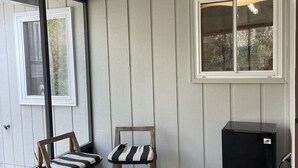 Een kamer met een houten vloer, twee stoelen met gestreepte kussens, een zwarte koelkast en een raam met uitzicht op bomen.