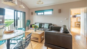 A modern living room with a sofa, coffee table, and dining table. There is a large window with a view of greenery outside. - Buttercup lodge - sleeps 4/5 with hot tub & countryside views (Cheddar)