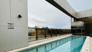 En moderne indendørs swimmingpool med klart, blåt vand, omgivet af en flisbelagt platform og et glasgelænder.
