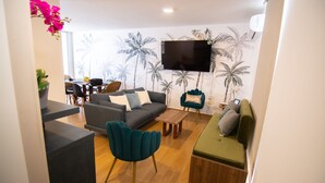 Un salon moderne avec un canapé gris, un fauteuil turquoise et un pouf vert. Un téléviseur à écran plat est fixé au mur, et il y a un coin repas avec des chaises et une table.