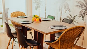 Une table à manger avec un saladier de fruits, des chaises noires en osier et un papier peint aux motifs tropicaux.