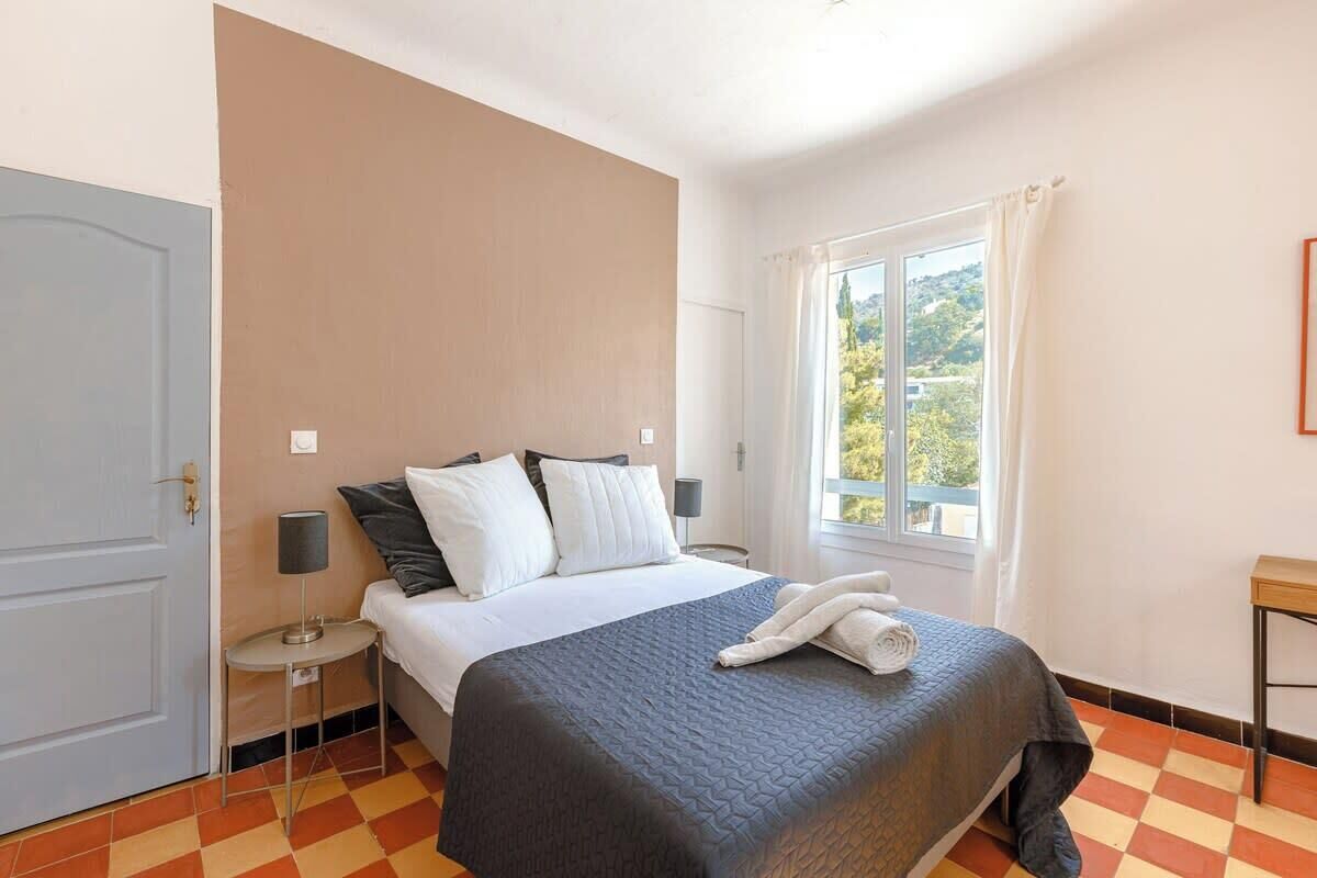 Ein Hotelzimmer mit Bett, Nachttisch, Lampe, einem Fenster mit Vorhängen und Blick auf Grünfläche.