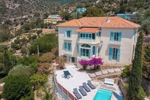 A two-story house with a tiled roof, a swimming pool, and a garden with flowering plants.
