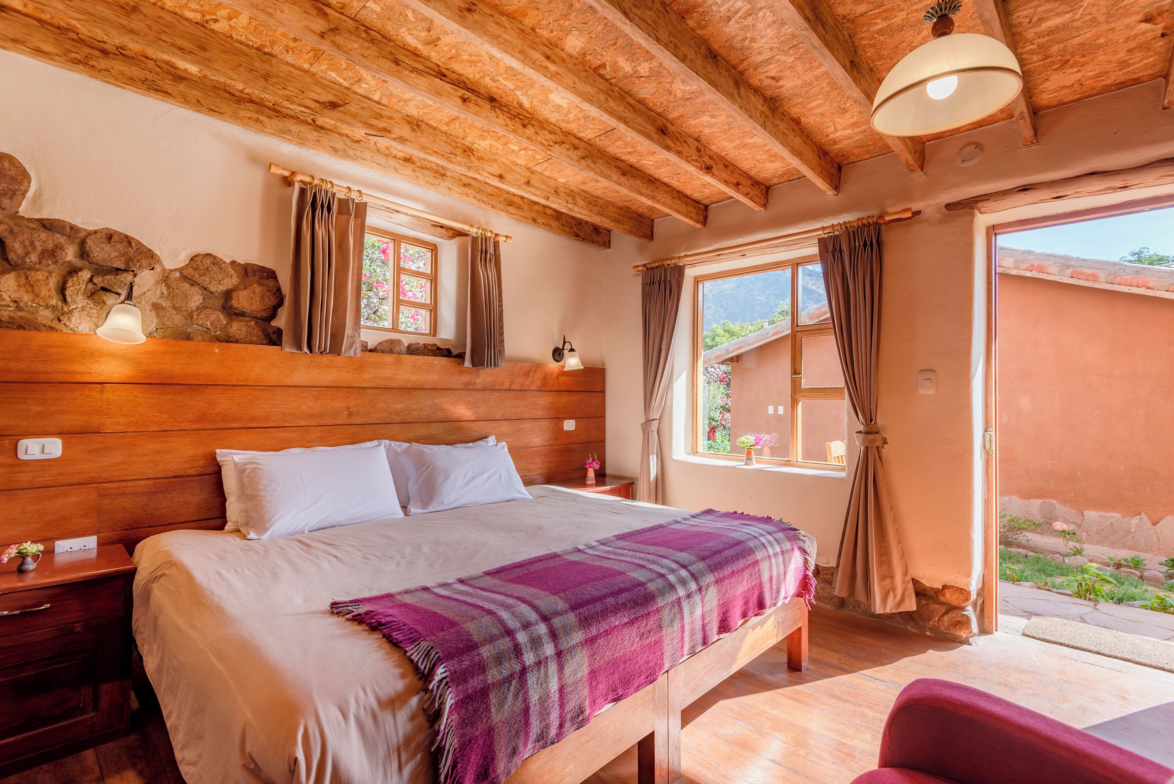 A bedroom with a wooden headboard, a large bed with a purple blanket, a nightstand with a lamp, and a window with curtains.