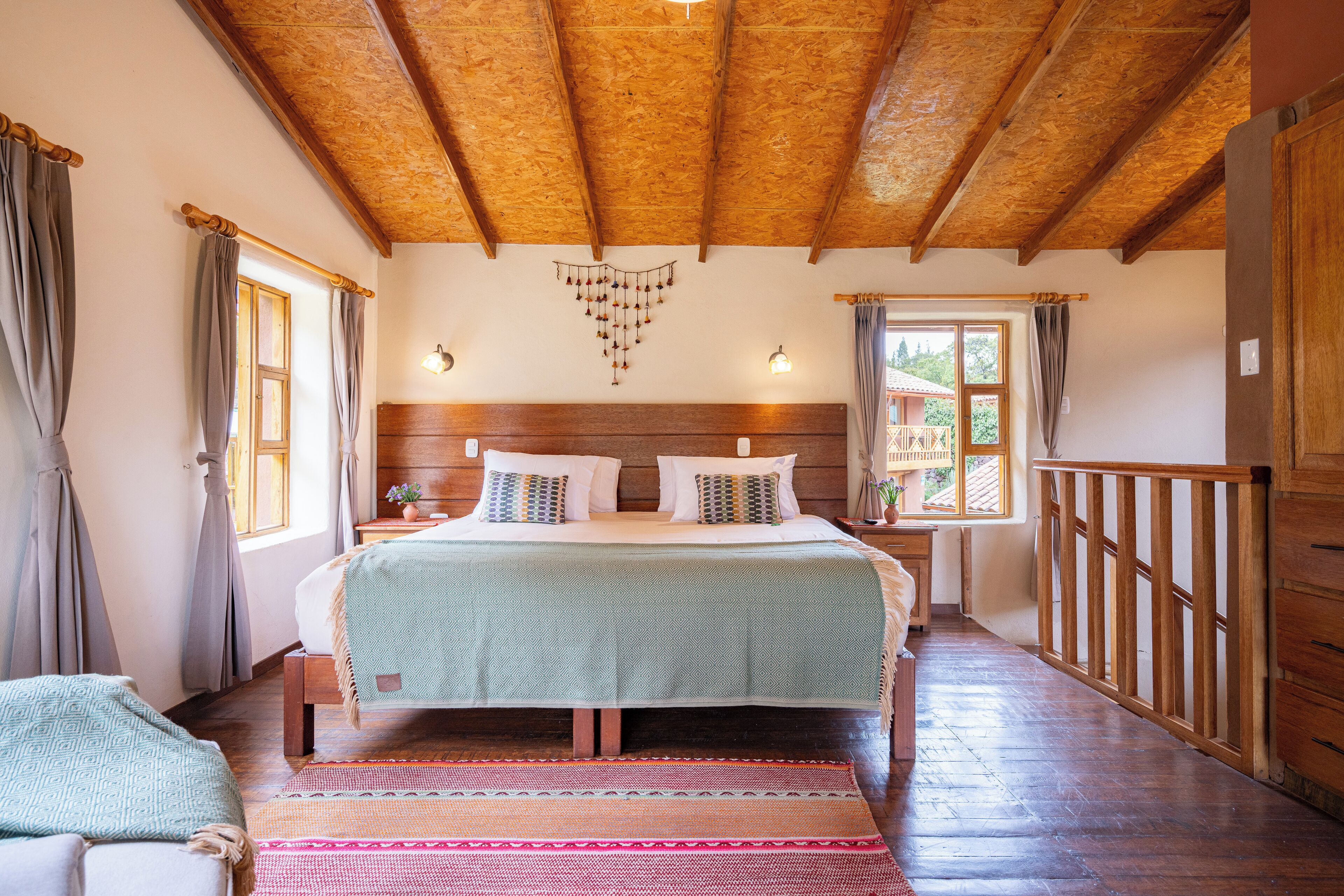 Un dormitorio con techo de madera, una cama con colcha verde, dos mesitas de noche y una ventana con cortinas.