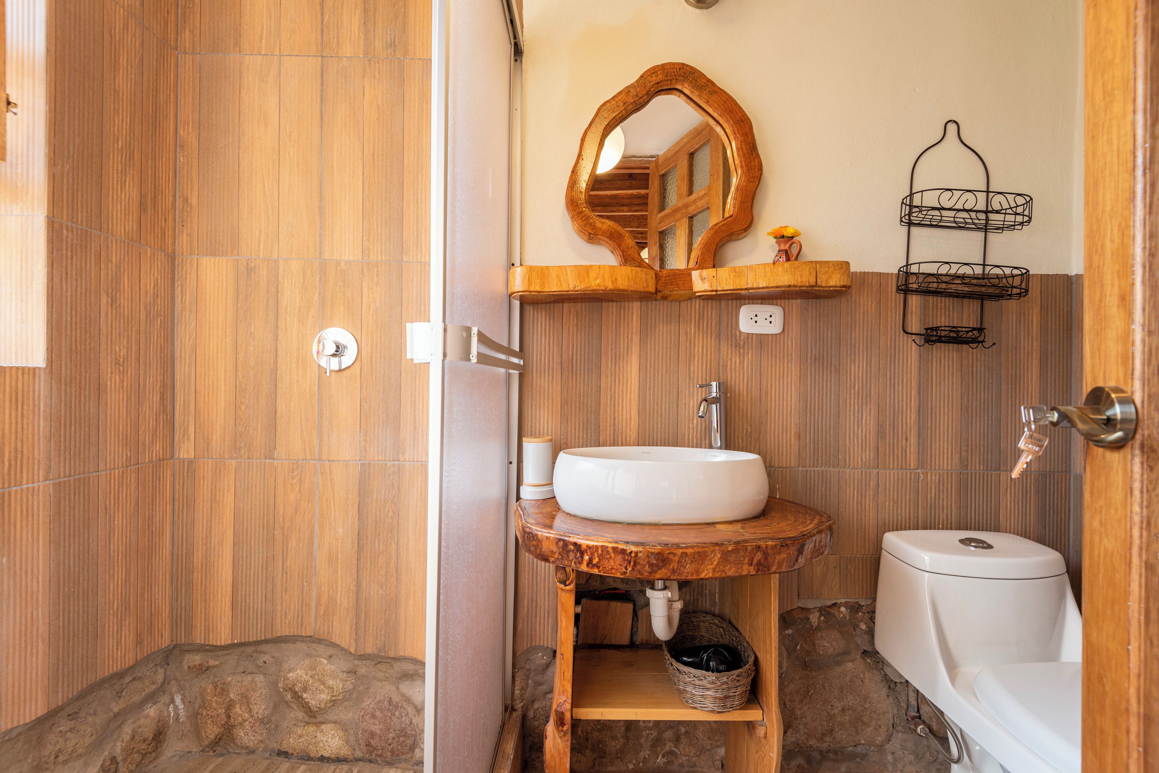 A bathroom with a wooden vanity, a white sink, a mirror, a toilet, and a small shelf with a basket.