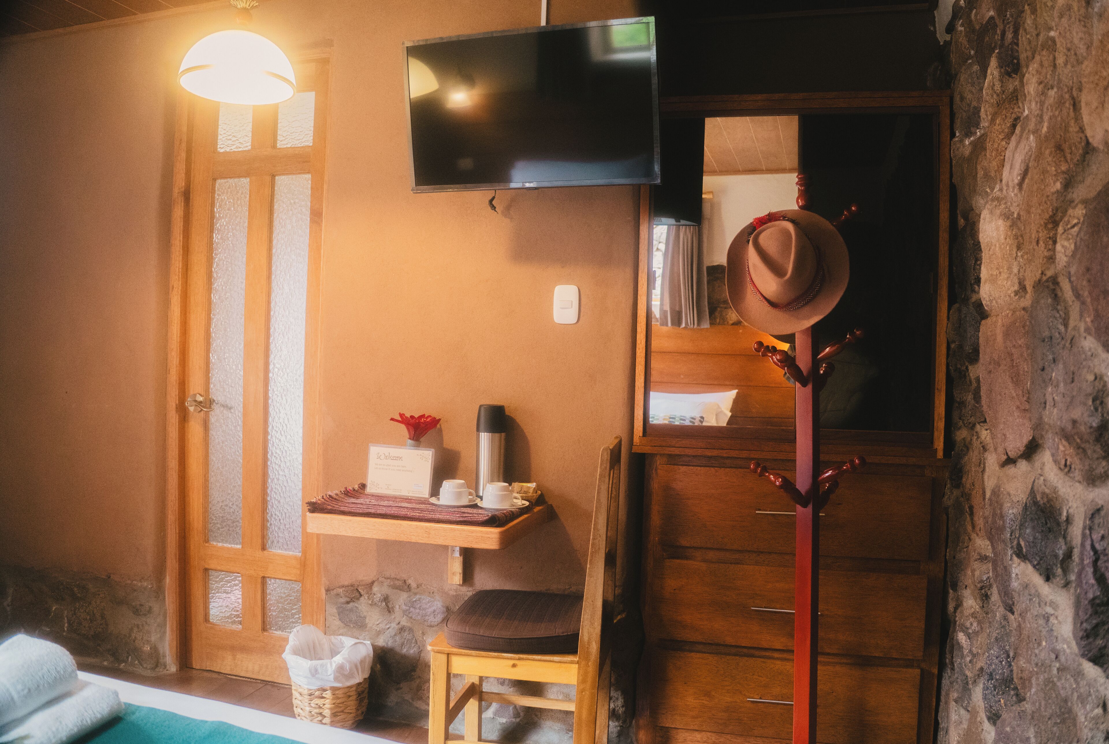 Um quarto acolhedor com uma cadeira de madeira, uma mesa pequena com xícaras e um termo, uma televisão fixada na parede e uma parede de pedra.