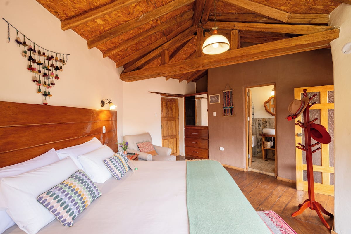 A bedroom with a wooden ceiling, a bed with multiple pillows, a hanging decoration, and a wooden door with a hat rack.