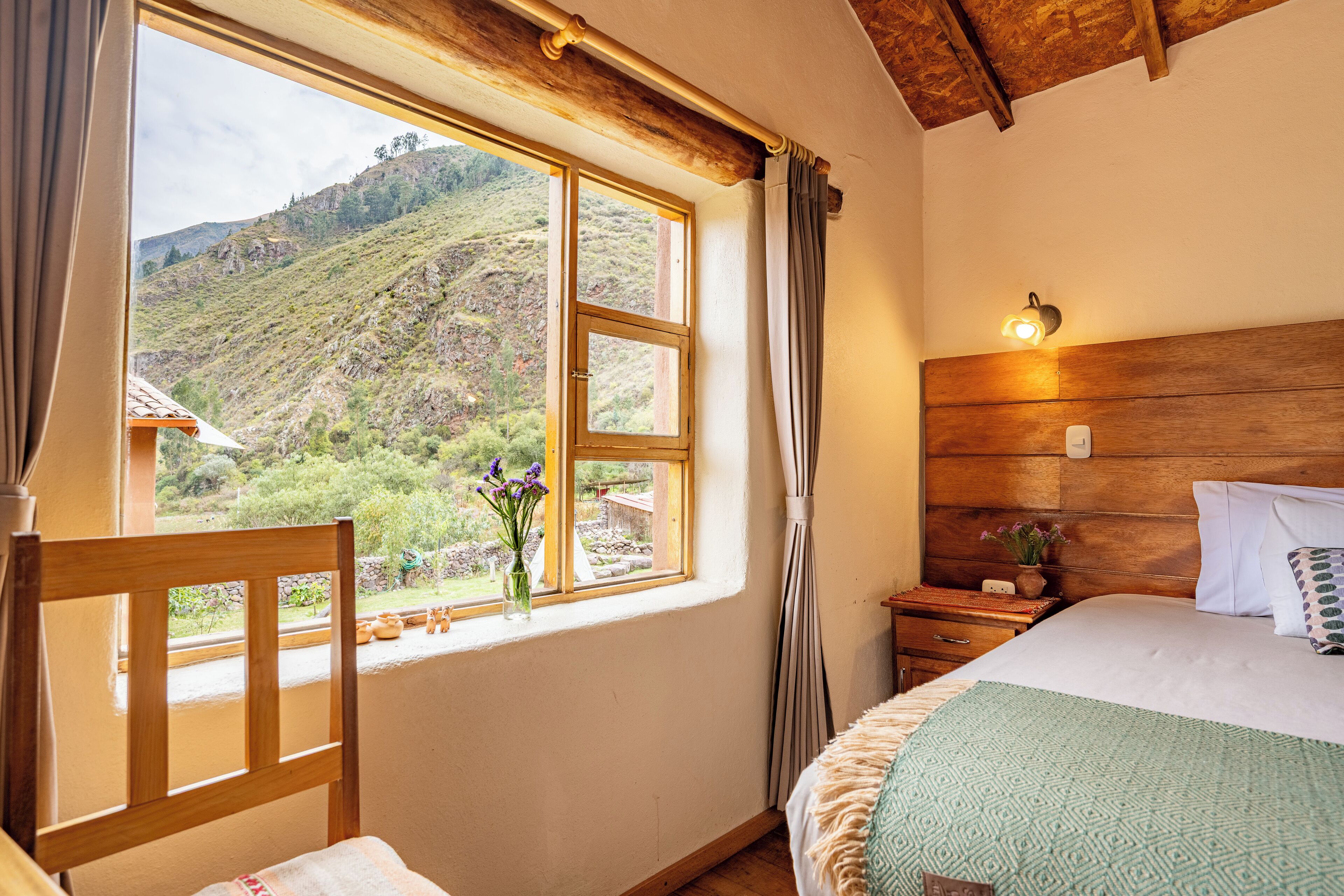A bedroom with a large window, wooden headboard, bedside table, and a bed with a patterned blanket.