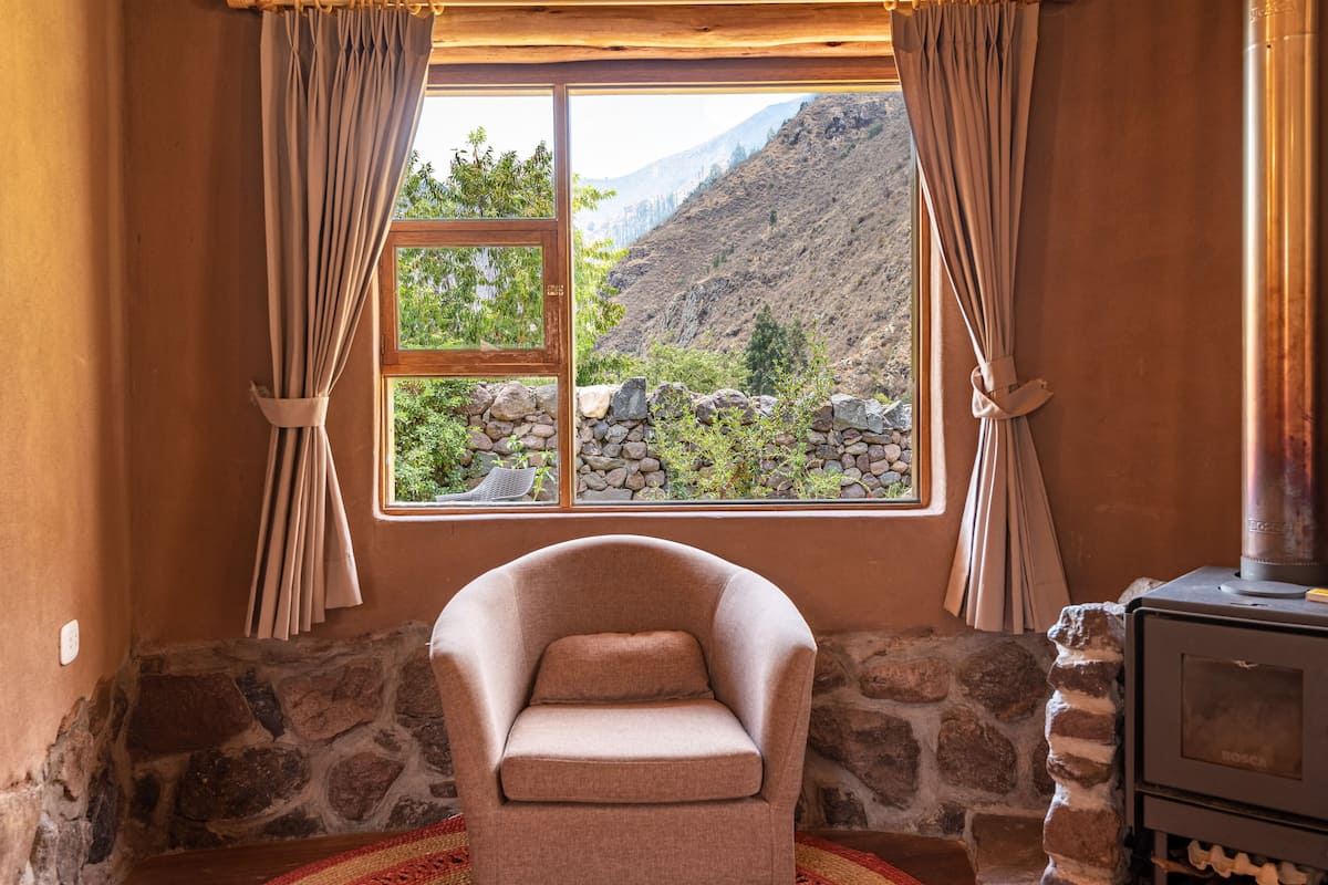 A cozy room with a stone wall, a window with curtains, a chair, and a wood-burning stove.