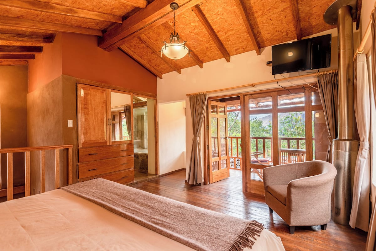A spacious bedroom with a large bed, wooden ceiling beams, a television, and a view of the outdoors through a sliding glass door.