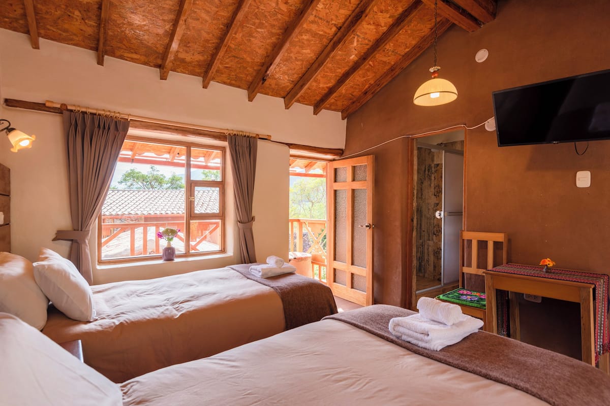 A hotel room with two beds, a wooden ceiling, a TV, and a window with curtains.