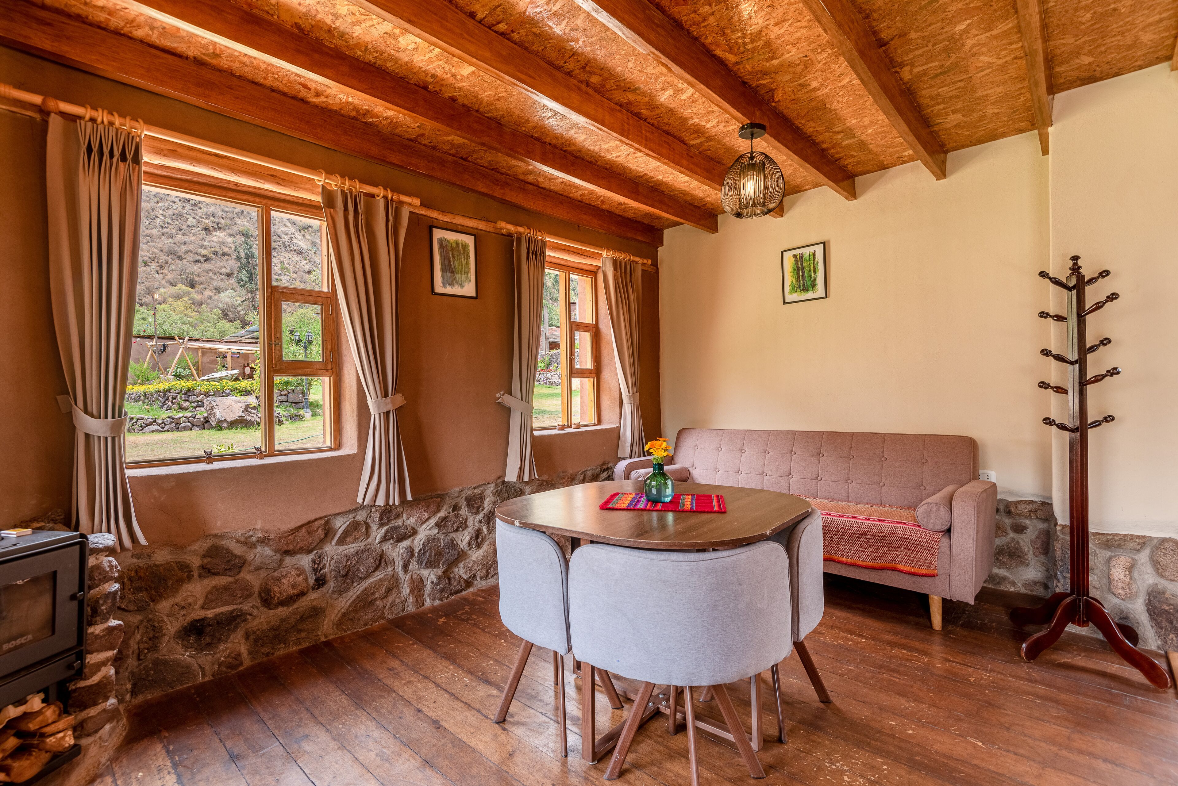 A cozy room with a stone wall, wooden ceiling beams, a round wooden table with two chairs, a pink sofa, and a fireplace.