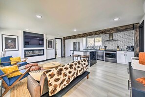 Een moderne woonkamer met een bank, stoelen en een open haard. De keuken heeft een groot eiland, roestvrijstalen apparatuur en een decoratieve achterwand.