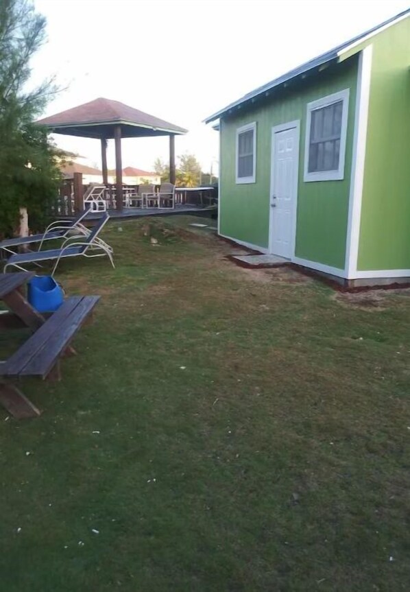 A small, single-story building with a green exterior and a white door. There is a wooden picnic table with benches and a blue container nearby. - A Hidden Gem (Nassau, Bahamas)