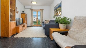 Una sala de estar con sofá, televisión, mueble de madera, planta y vista al exterior a través de un ventanal.